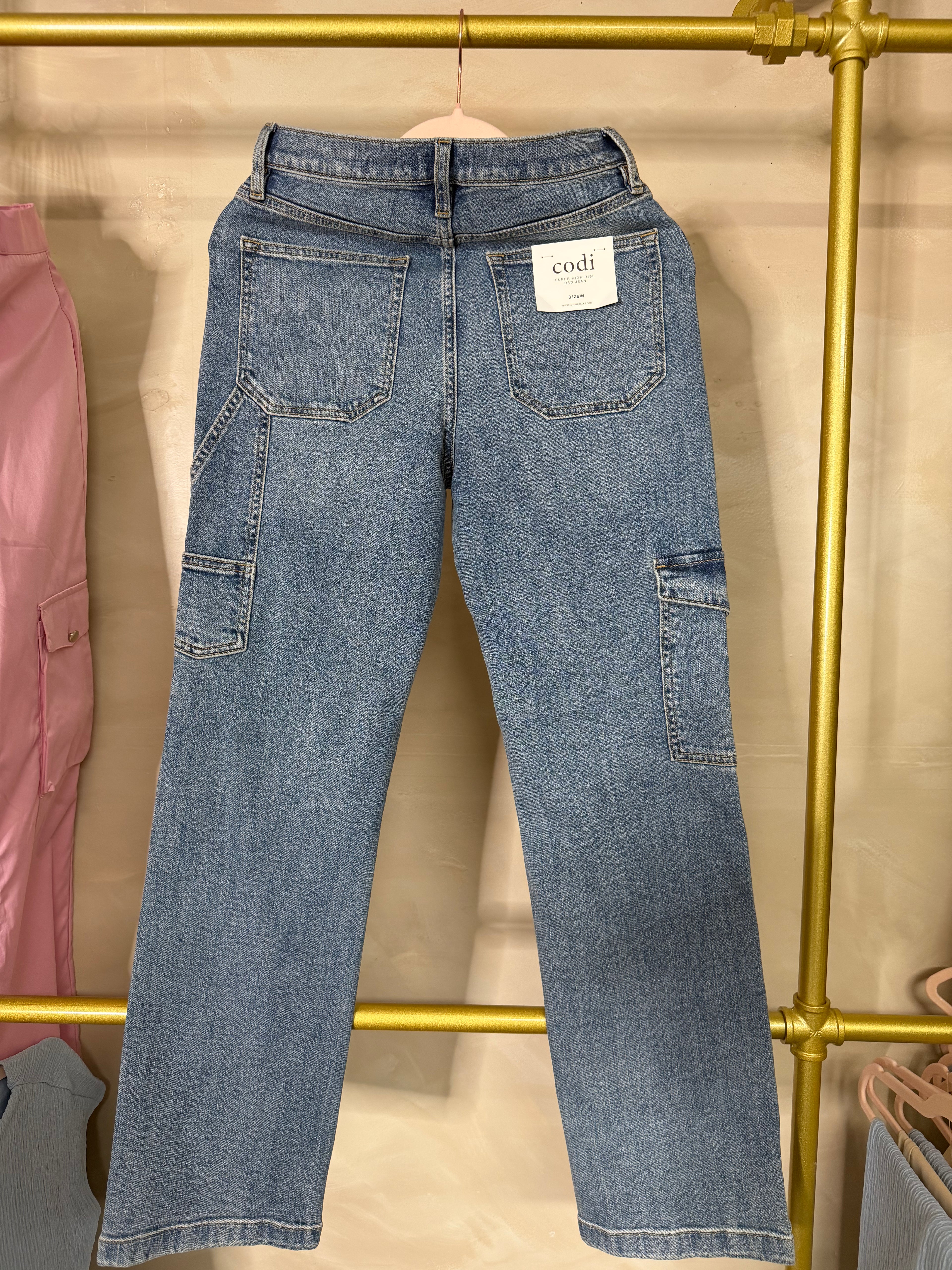 Light blue denim jeans with cargo pockets, shown from the back hanging on a gold clothing rack.